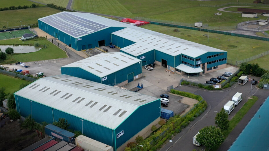 Aerial view of a large industrial complex with multiple blue warehouses and a parking area. Surrounding green fields are visible, along with some vehicles and a small body of water nearby.