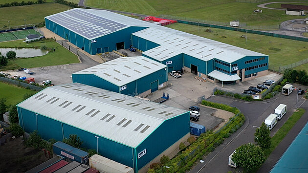 Aerial view of a large industrial complex with multiple blue warehouses and a parking area. Surrounding green fields are visible, along with some vehicles and a small body of water nearby.