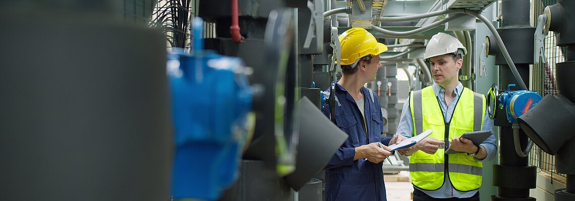 Two workers in safety gear discuss documents near machinery in an industrial setting.