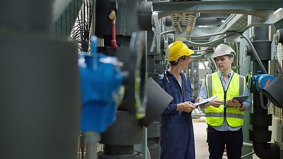Two workers in safety gear discuss documents near machinery in an industrial setting.