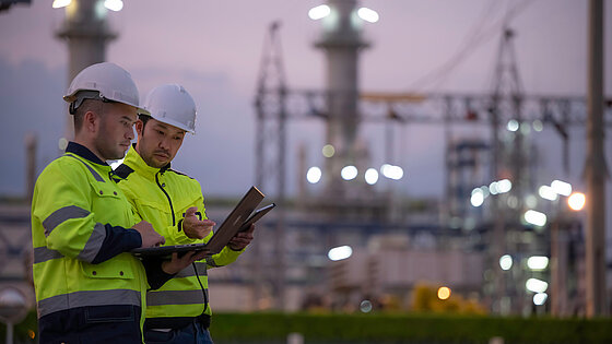 Two workers in safety gear review a document at an industrial site.