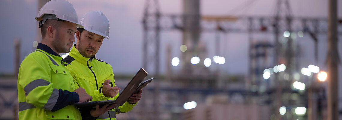 Two workers in safety gear review a document at an industrial site.