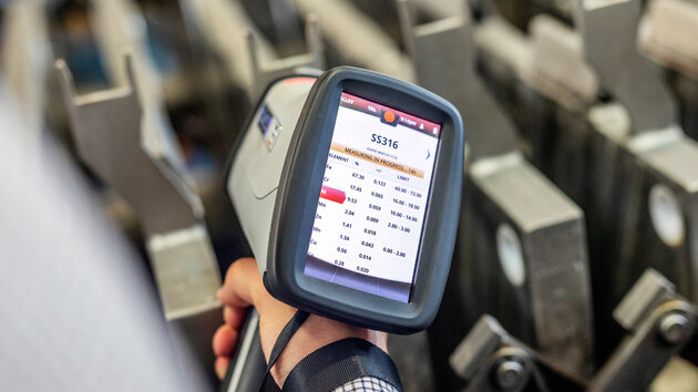A hand holds a scanning device displaying a data screen with numbers and labels. In the background, there are metal racks or structures. The focus is on the scanning device in the foreground.