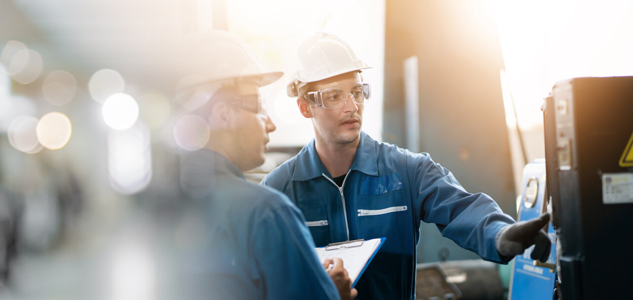 Two workers in safety helmets and glasses are discussing equipment details in a workshop. One is holding a clipboard and pointing at a machine, while the other is listening attentively. The environment is bright with industrial elements in the background.