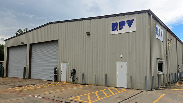 A large, gray metal building with a logo that reads "RPV" displayed prominently on the front. The building features two large garage doors, a pedestrian entrance, and a parking area in front. A white vehicle is parked nearby on a paved surface.