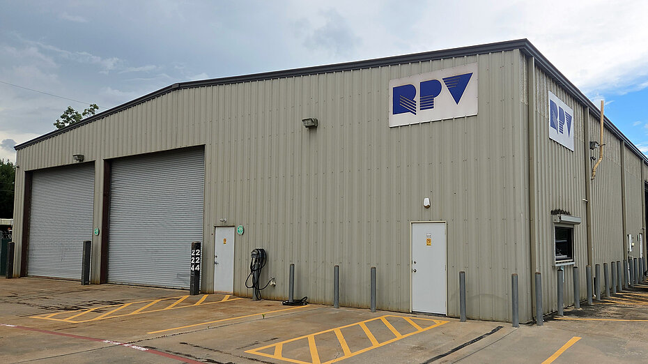 A large, gray metal building with a logo that reads "RPV" displayed prominently on the front. The building features two large garage doors, a pedestrian entrance, and a parking area in front. A white vehicle is parked nearby on a paved surface.