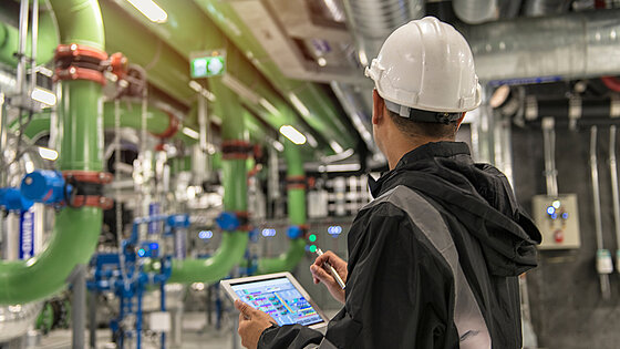 A person in a hard hat uses a tablet near large green pipes and blue valves.