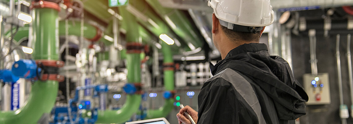 A person in a hard hat uses a tablet near large green pipes and blue valves.