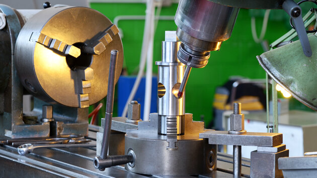 A close-up view of a machining setup featuring a metal lathe and a milling machine. The foreground shows a metal piece being machined, with tools and fixtures positioned around it. The background includes a green wall and various workshop equipment.