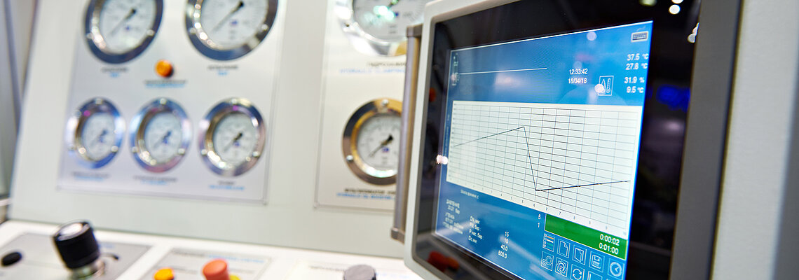 A control panel featuring multiple pressure gauges and a digital touchscreen display. The display shows a graph with readings and various control knobs and buttons in the foreground. The panel is designed for monitoring and operational control, likely in an industrial or technical setting.