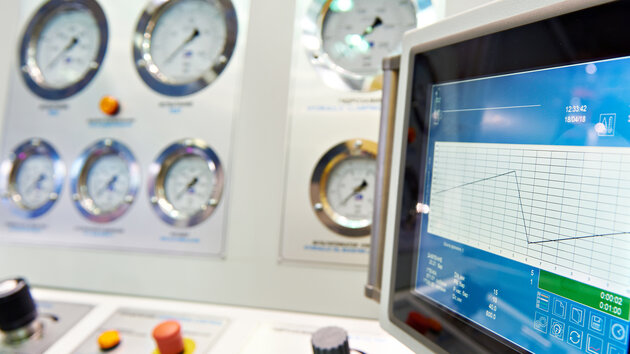 A control panel featuring multiple pressure gauges and a digital touchscreen display. The display shows a graph with readings and various control knobs and buttons in the foreground. The panel is designed for monitoring and operational control, likely in an industrial or technical setting.