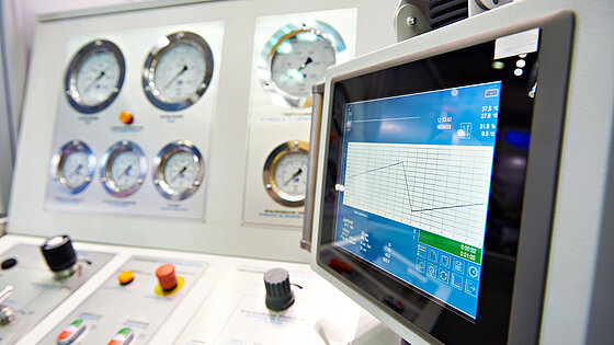 A control panel featuring multiple pressure gauges and a digital touchscreen display. The display shows a graph with readings and various control knobs and buttons in the foreground. The panel is designed for monitoring and operational control, likely in an industrial or technical setting.
